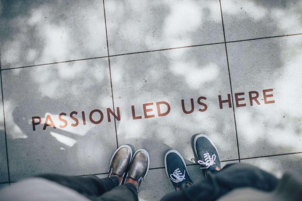 This image shows two individuals standing side by side, viewed from above, on a paved sidewalk with the motivational phrase “Passion Led Us Here” inscribed on the surface. The scene is softly lit with natural shadows, symbolizing purpose, unity, and the journey driven by passion.