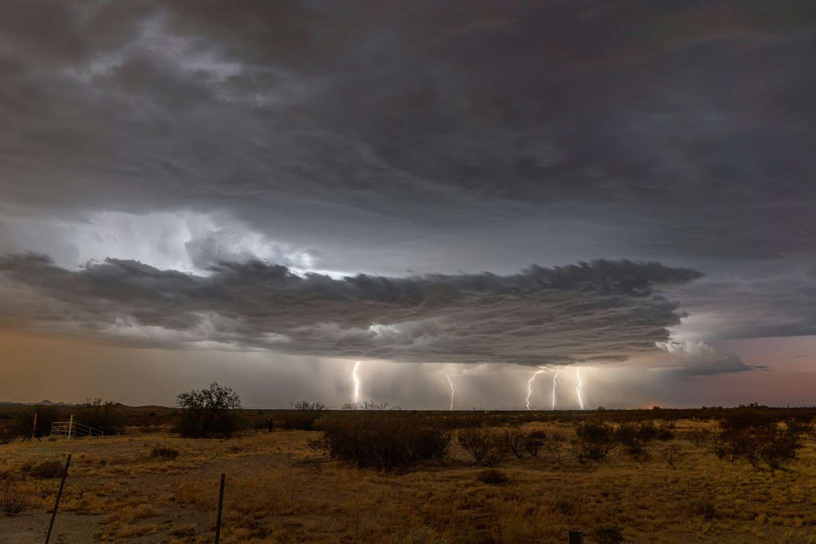 Monsoon Preparedness Arizona 2025-A dramatic desert landscape under a dark, stormy sky. Thick, layered clouds dominate the scene as multiple lightning bolts strike the ground in the distance. Sparse desert vegetation and a dry, brown field are visible in the foreground.